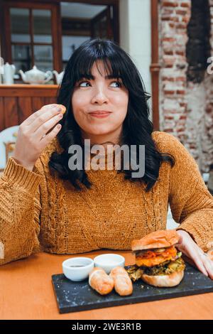 Vertikales Bild der jungen Latina mit schwarzen Haaren und braunen Kleidern, glückliches Sitzen im Restaurant und Tequenos, traditionelles venezolanisches Essen, mit Hambu Stockfoto