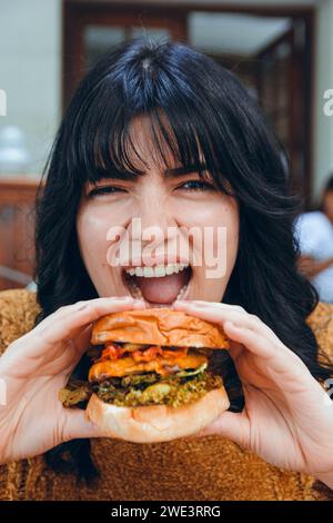 Vertikales Nahbild einer hungrigen kaukasischen Frau, die großen Rindfleischburger mit Käse und Gemüse hält und isst, Bild für soziale Netzwerke und schnell Stockfoto