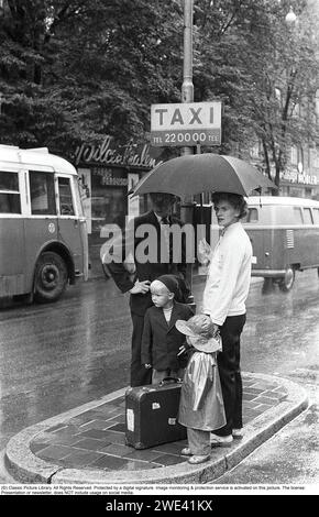 Stockholm in den 1950er Jahren Es ist ein regnerischer Tag im Zentrum Stockholms. Ein junges Paar steht unter einem Schirm an einem Taxistand und wartet auf ein Taxi, aber bei regnerischem Wetter kann es eine lange Wartezeit unter dem Schirm für das junge Paar sein. Nur eines der Kinder trägt Regenkleidung. Stockholm 1953. Kristoffersson Ref. 2A-12 cv26-3 Stockfoto