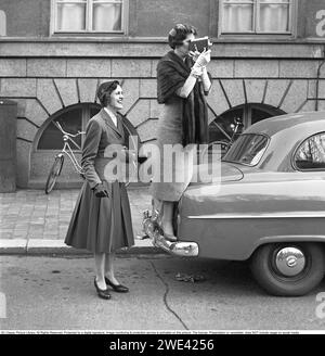 In den 1950er Jahren Eine ältere Frau mit einer Filmkamera stand auf dem Stoßfänger eines Autos, um einen besseren Blick auf das zu bekommen, was sie gerade filmt. Eine jüngere Frau steht daneben und sieht glücklich aus. Sie sind Verwandte eines der kürzlich abgeschlossenen Studenten, die feiern, dass sie ihre Immatrikulationsprüfung bestanden haben und eine Stockholmer Straße hinunterlaufen. 1954. Svahn Ref. SVA2 Stockfoto