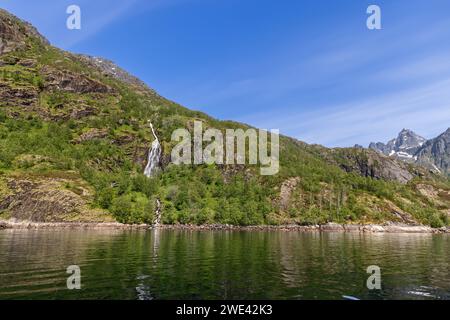 Kristallklares Wasser stürzt von einer hohen Klippe in den ruhigen Fjord, umgeben von den lebhaften Grünflächen eines norwegischen Sommers im Trollfjord, Lofoten Stockfoto