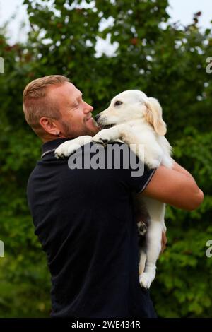 Ein zufriedener Mann genießt einen warmen Sommertag im Freien, während er liebevoll einen jungen, flauschigen goldenen Retriever-Welpen in den Armen hält. Stockfoto