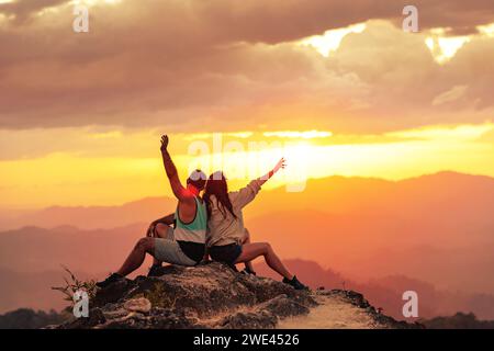 Glückliches Paar genießt den Sonnenuntergang und sitzt mit offenen Armen auf dem Gipfel des Berges Stockfoto