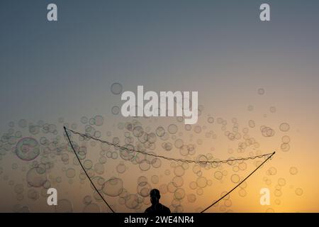 Ein Mann in der Silhouette macht viele Seifenblasen mit einem Seifenblasenstab, sie schweben am orangefarbenen Himmel bei Sonnenuntergang Stockfoto