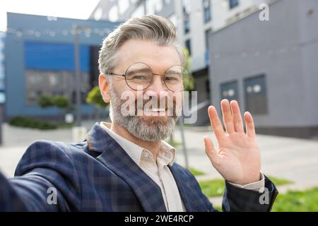 Erfahrener Geschäftsmann in einem eleganten Anzug, der vor einem Büro hallo winkt und Professionalität und Freundlichkeit verkörpert. Stockfoto