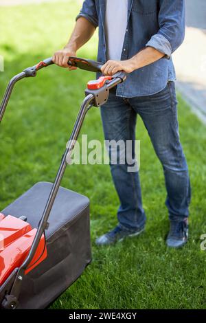 Nicht erkennbarer männlicher Gärtner, der modernen Rasenmäher drückt, während er auf Gras geht. Über dem Blick auf den Handwerker aus Denim, der sich mit dem Rasentrimmer im Freien um den Rasen kümmert. Konzept der Gartenausrüstung. Stockfoto