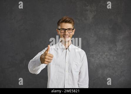 Glücklicher junger Mann in einer Brille, der die Daumen hoch hält und isoliert auf grauem Hintergrund steht Stockfoto