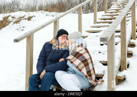 Glückliches älteres Paar, das im Winter auf einer Holztreppe sitzt und mit einer Decke bedeckt ist. Glückskonzept. Stockfoto