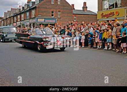 Watfords jährliche Pfsun-Karnevalsparade entlang der St Albans Road, Watford, Hertfordshire, England, Vereinigtes Königreich, 1967. ein Nachbau von Batmobile nahm Teil – das Auto erschien 1939 im DC-Comic Batman und in TV-Serien der späten 1960er Jahre – es zeigte Batman (Bruce Wayne) und sein Freund Robin (ein maskierter Robin befindet sich auf dem Beifahrersitz) benutzte ihr Fahrzeug, um Verbrechen zu verhindern und Bösewichte zu fangen. Ein Watford Observer Bedford Van folgt dem Batmobil – einem Vintage-Foto aus den 1960er Jahren. Stockfoto