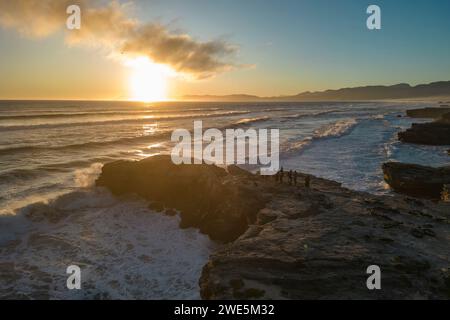 Aus der Vogelperspektive sehen Sie Menschen, die auf einem Felsvorsprung mit fetten Fahrrädern stehen, während Wellen bei Sonnenuntergang auf die Küste und den Strand im Walker Bay Nature Reserv brechen Stockfoto