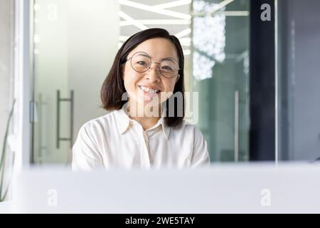 Porträt einer zufriedenen asiatischen Frau in runder transparenter Brille, die die Kamera mit einem zahnlosen Lächeln am Arbeitsplatz betrachtet. Erfolgreicher Projektmanager mit kurzen Haarschnitten, der einen produktiven, geschäftigen Tag im Büro genießt. Stockfoto