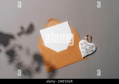 Umschlag mit leerer Karte, weißem Vintage-Herz und Blumenschatten auf grauem Hintergrund. Draufsicht, flach, Modell. Stockfoto