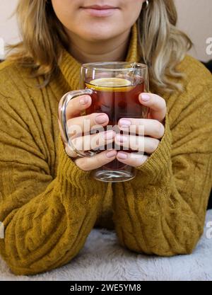 Nahaufnahme von Frauenhänden, die eine Glasschale mit heißem Tee mit Zitrone und Honig halten Stockfoto