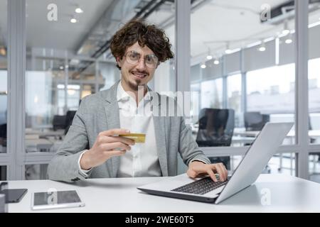 Ein selbstbewusster männlicher Büroangestellter ist an Online-Transaktionen beteiligt und zeigt an seinem lichtdurchfluteten Arbeitsplatz Fähigkeiten und positive Emotionen. Stockfoto