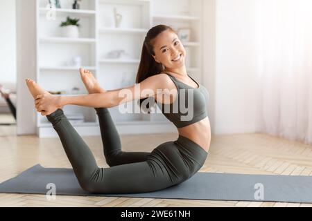 Fitte junge asiatische Frau, die zu Hause eine anspruchsvolle Yoga-Pose macht und lächelt Stockfoto