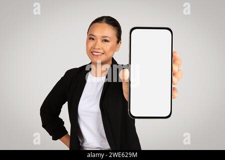 Lächelnde asiatische Geschäftsfrau, die ein Smartphone mit einem leeren Bildschirm in Richtung Kamera präsentiert Stockfoto