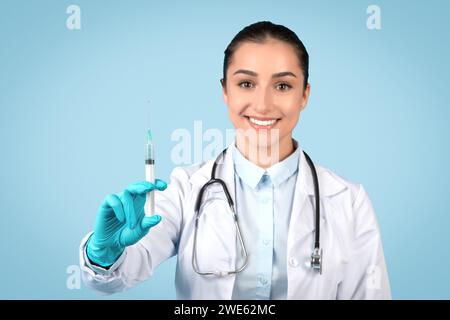 Lächelnde europäische Ärztin trägt sterile Handschuhe, hält medizinische Spritze, blauer Atelierhintergrund Stockfoto