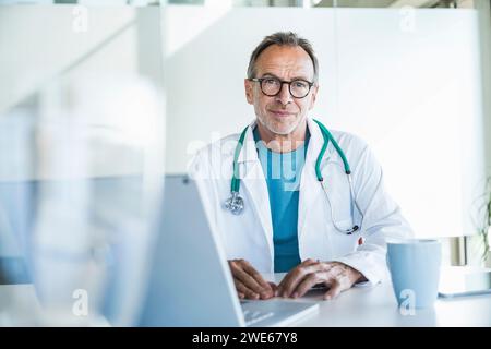 Lächelnder Seniorarzt sitzt am Schreibtisch Stockfoto