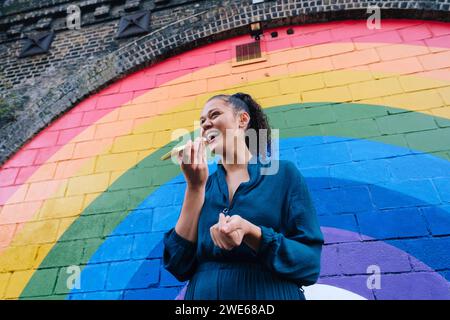 Glückliche junge Frau, die Voicemail über das Smartphone vor der Regenbogenwand schickt Stockfoto