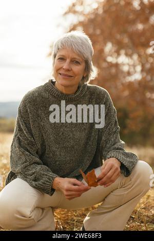 Lächelnde reife Frau, die Blatt hält und sich auf dem Feld hockt Stockfoto