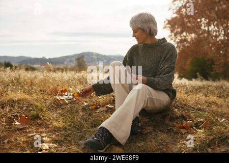 Reife Frau, die auf Gras sitzt und Blätter auf dem Feld hält Stockfoto