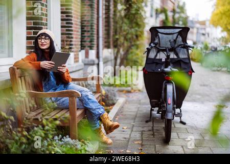 Lächelnde Frau, die auf einer Bank sitzt und einen Tablet-PC in der Nähe eines Lastenfahrrads hat Stockfoto