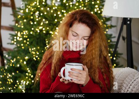 Glückliche Rothaarige Frau, die zu Hause eine Kaffeetasse vor dem Weihnachtsbaum hält Stockfoto