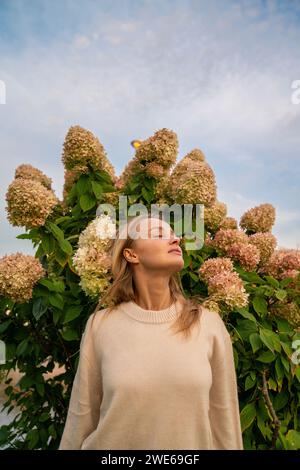 Junge Frau, die mit geschlossenen Augen vor blühender Pflanze unter dem Himmel steht Stockfoto