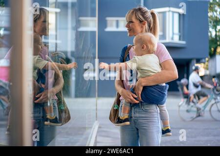 Mutter macht Schaufensterbummel mit Sohn im Babytragetuch auf der Straße Stockfoto