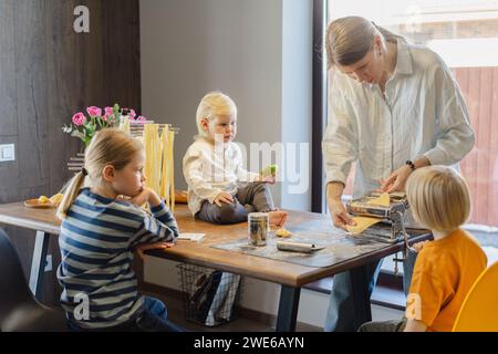 Frau, die Teig in der Pasta-Maschine schneidet, mit Familie, die am Esstisch sitzt Stockfoto