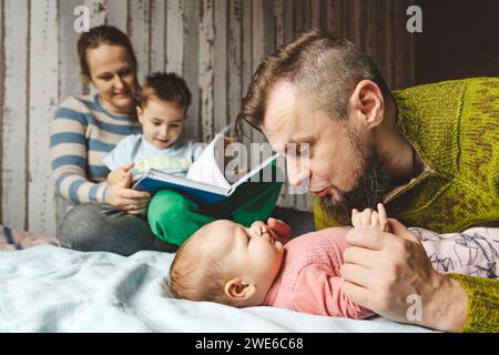 Vater, der das Baby umschließt, Tochter und Mutter liest Buch mit Sohn zu Hause Stockfoto