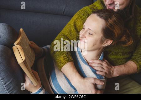 Lächelnde Frau, die auf dem Schoß des Mannes liegt und zu Hause Buch liest Stockfoto