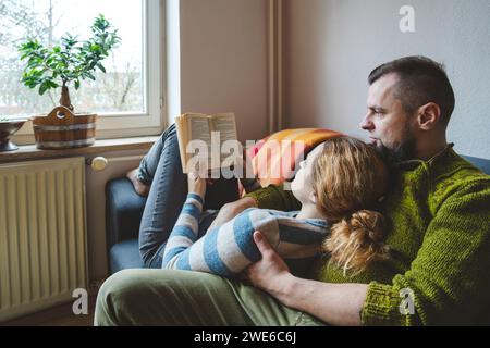 Frau, die auf dem Schoß des Mannes liegt und zu Hause Buch liest Stockfoto