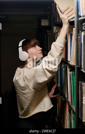 Junger Schüler, der kabellose Kopfhörer trägt und Buch aus dem obersten Regal der Bibliothek nimmt Stockfoto