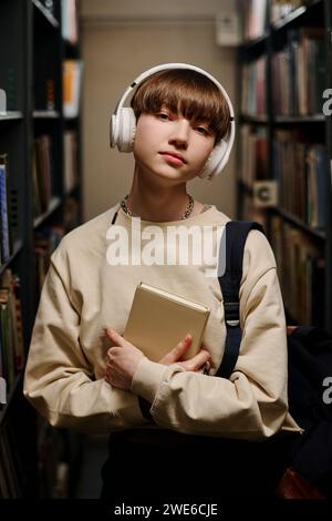 Junger Student, der kabellose Kopfhörer trägt und ein Buch hält Stockfoto
