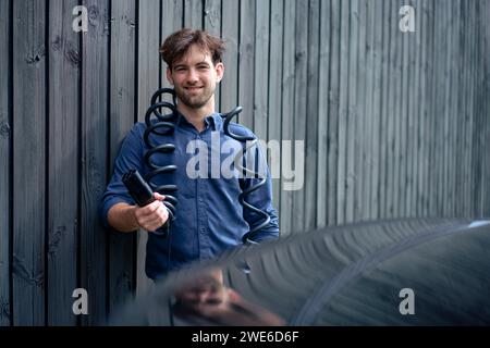 Lächelnder Mann mit elektrischem Ladestecker des Autos, der vor der Wand steht Stockfoto