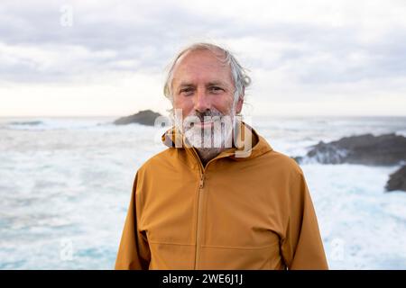 Lächelnder Seniorenmann, der Jacke trägt und in der Nähe des Meeres steht Stockfoto