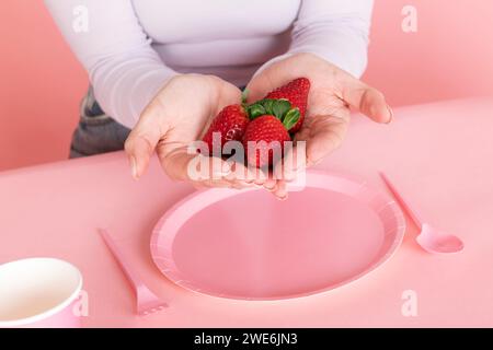 Frau, die Erdbeeren in der Hand hält, über einem pinkfarbenen leeren Teller Stockfoto