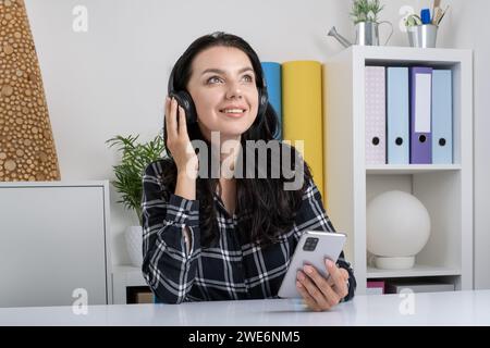 Musik hören, während Sie kabellose Kopfhörer tragen. Junge glückliche, lächelnde junge Frau im Büro. Stockfoto