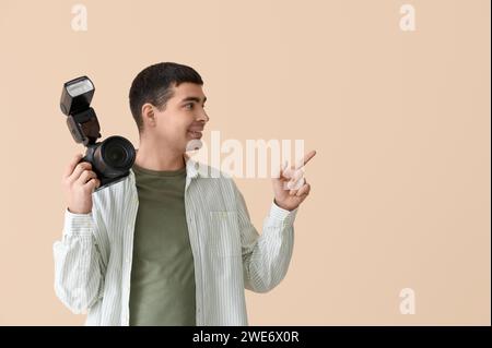 Junger männlicher Fotograf mit professioneller Kamera, der auf etwas auf beigefarbenem Hintergrund zeigt Stockfoto