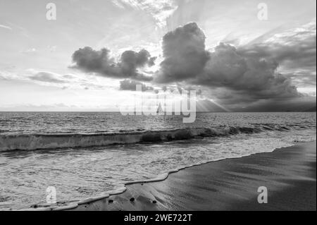 Ein Segelboot segelt entlang des Ozeans mit Sonnenstrahlen, die durch die Wolken in Schwarz und weiß brechen Stockfoto