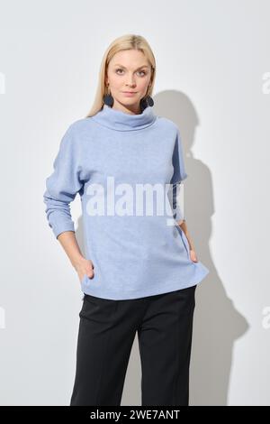 Studio-Porträt einer klassischen blonden Frau in blauem Pullover und schwarzer Hose mit Händen in Taschen Stockfoto