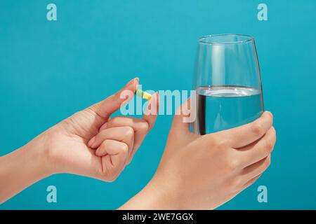 Die minimale Szene einer gelb-grünen Kapsel wird von einem Handmodell gehalten, einem mit Wasser gefüllten Glas in einer anderen Hand. Vitamin kann die Lebensqualität erhöhen Stockfoto