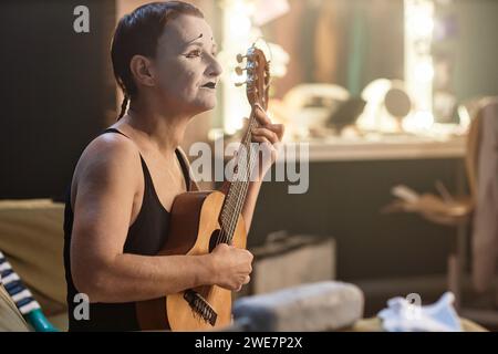Seitliches Porträt einer Mime-Darstellerin, die Ukulele spielt, während sie Backstage-Proben, Kopierraum Stockfoto