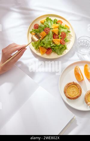 Das Handmodell hält eine goldene Gabel, um eine orangene Scheibe auf die runde Schale zu nehmen. Ein Glas Wasser wird angezeigt. Whitepaper ohne Text Stockfoto