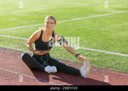 Passen Sie weibliche Athletin, die auf einer Sportstrecke sitzt und ihr Bein ausdehnt und ihr Telefon, Kopfhörer in die Trainingspause benutzt. Stockfoto