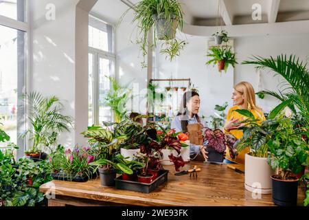 Junge asiatische Verkäuferin in Schürze, die mit weiblichen Kunden im Blumenladen spricht. Stockfoto