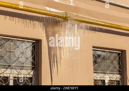 Große Eiszapfen auf dem Dach eines Stadthauses an einem sonnigen Wintertag unter Taue. Eiszapfen hängen an einer Gasleitung an der Wand eines Wohnhauses. Stockfoto