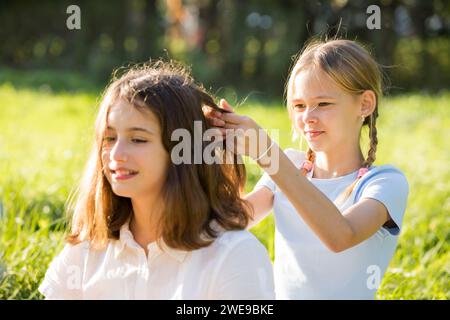 Zwei Teenager verbringen Zeit auf grünem Gras im Park, Zöpfe und Schwänze für einander, genießen Sommer und Urlaub Stockfoto
