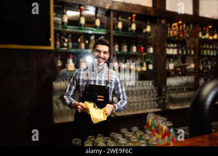 Junger, glücklicher Barkeeper, der das Trinkglas putzt. Ein gutaussehender Barkeeper poliert ein Glas Wein. Das Konzept des Service im Coffee Shop. Stockfoto